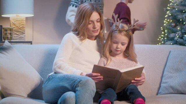 Beautiful Caucasian mother with her daughter reading book while sitting on sofa at home. Positive family having recreation on winter holidays. In background father and son decorating Christmas tree.