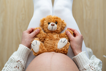 Close-up high angle shot of a cute stuffed bear sitting on mothers naked, round belly. Last month of pregnancy - week 39. White background. Bright shot.