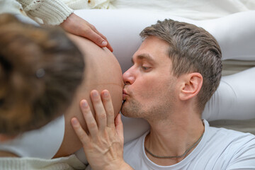 Close-up of the parents lying on the bed, with the father holding and kissing the naked round baby bump of the pregnant mother. Last month of pregnancy - week 39. White background. Bright shot. - obrazy, fototapety, plakaty