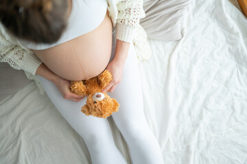 Pregnant mother sits on the bed and holds a cute stuffed bear to her round baby bump. Last month of...