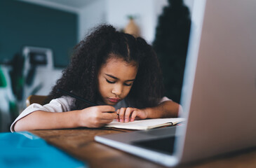 Naklejka premium Focused ethnic girl writing in notepad while sitting at table