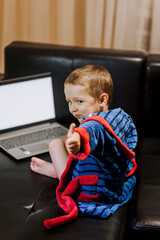 Fototapeta premium A small handsome smiling boy, a child in pajamas, sits on the sofa in the room, shows like near the laptop, playing games, watching interesting movies via the Internet. Childhood concept, copy space.