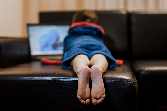 A Boy Child In Pajamas Lies On A Black Leather Sofa, Watching A Movie On A Laptop Online Via The Internet. Legs, Feet Close-up. Happy Childhood, Lifestyle.