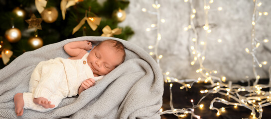 New Year Christmas holidays scene - cute sleeping newborn baby boy in a beige knitted jumpsuit onesie lying on blanket with Christmas festive tree gold toys and lights garland. Infant,Nursery - obrazy, fototapety, plakaty
