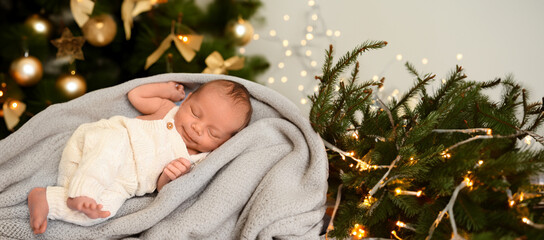 New Year Christmas holidays scene - cute sleeping newborn baby boy in a beige knitted jumpsuit onesie lying on blanket with Christmas festive tree gold toys and lights garland. Infant,Nursery