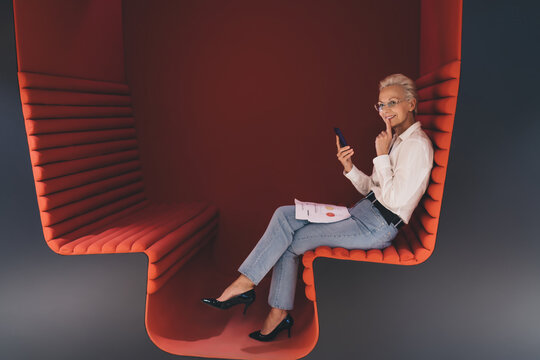 Thoughtful senior Caucasian businesswoman using smartphone, sitting on a modern red couch, with documents, in an office setting