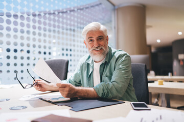 Cheerful senior Caucasian man with a white beard, holding glasses and paperwork in a bright office space, exuding confidence and approachability