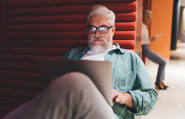Focused senior man with grey beard and glasses working on a laptop in a modern lounge with red...