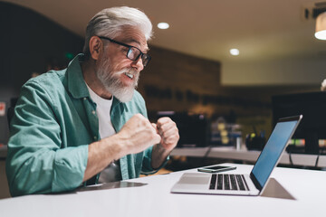 Elderly Caucasian man with glasses and a beard, showing excitement while using a laptop, possibly experiencing success or good news, in a casual office setting
