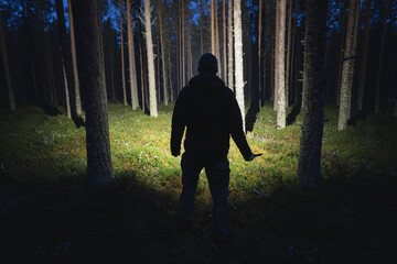 Silhouette of a man with a knife and a flashlight in a dark forest. - obrazy, fototapety, plakaty