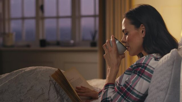 Woman at home lying in bed at night wearing pyjamas holding hot drink reading book - shot in slow motion