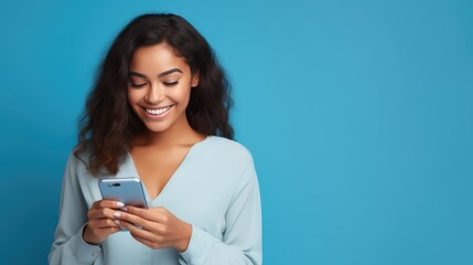 Fototapeta premium Radiant woman with dark skin in a blue shirt, beaming at her phone against a vivid blue backdrop, exuding joy