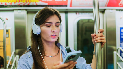 Happy young beautiful female passenger listening music via smart mobile phone in train. Smiling charming woman wearing wireless headphones while moving in tram. Big city lifestyle, transportation.