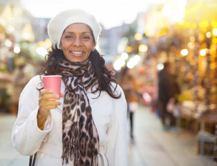 Portrait of smiling hispanic woman walking in colorful Christmas street market, holding paper cup of warm drink
