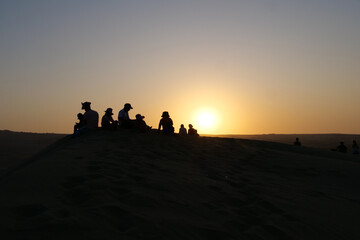Fototapeta na wymiar Black silhouettes of a group of people sitting on a dune at Huacachina oasis town, waiting for the sunset