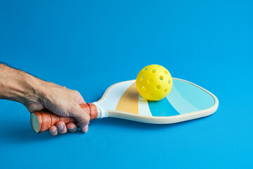 man's hand holding a pickleball paddle with a yellow ball on top on blue background