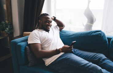 Cheerful black man browsing smartphone while sitting on sofa