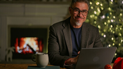 Happy smiling mid adult man using laptop computer at home, sitting at desk in Christmas decorated...