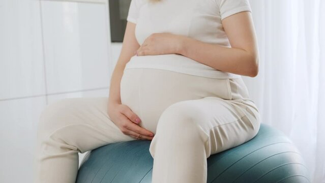 Pregnant woman slightly bouncing on rubber ball doing pregnancy gymnastics. Future mother gently touches growing belly in anticipation of baby crop body