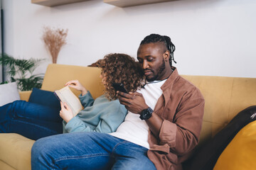 Focused multiethnic couple reading book and browsing smartphone while sitting together on sofa