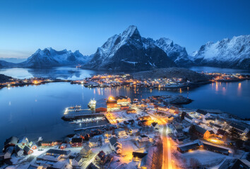 Aerial view of snowy village, islands, rorbu, city lights, blue sea, rocks and mountain at night in winter. Beautiful landscape with town, street illumination. Top view. Reine, Lofoten islands, Norway - obrazy, fototapety, plakaty