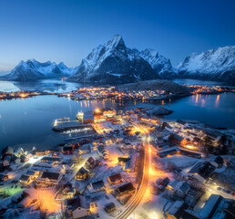 Aerial view of snowy village, islands, rorbu, city lights, blue sea, rocks and mountain at night in...