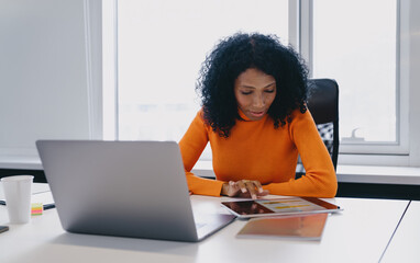 Engrossed African American cryptocurrency trader, analyzing live digital currency charts on tablet, with laptop beside in coworking, orange sweater symbolizing the energetic pulse of crypto market.