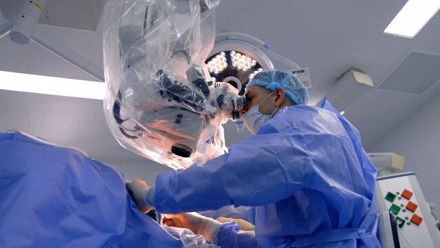 Concentrated male surgeon looking at microscope applies metal tool. Low angle view of the doctor working in surgery room.