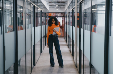 Black businesswoman in vibrant orange sweater and black jeans with a digital tablet in an office...