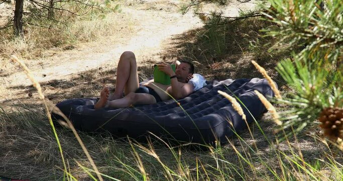 A man lies on an inflatable mattress in the woods on a sunny summer day, reading a book.