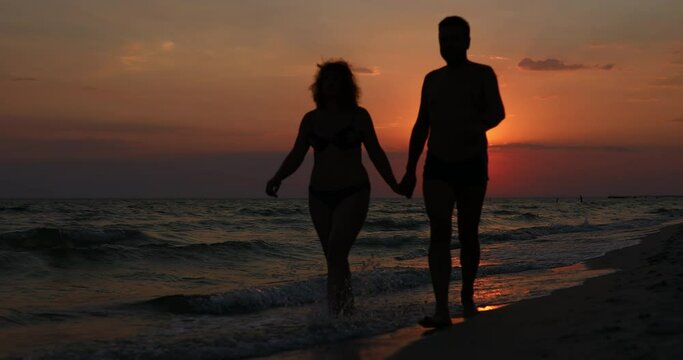 A man and a woman are walking along the seashore holding hands in front of a beautiful sunset.