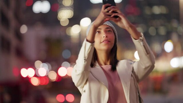 Trendy nightlife vlogger holding her phone and taking a photo of the city lights. Young woman admiring beautiful urban town, makes a video of the vibrant street view to share on her social media app