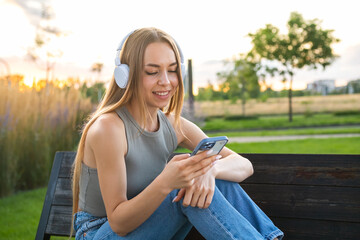 Carefree blonde woman listening to music using headphones and select music via smart phone sitting...