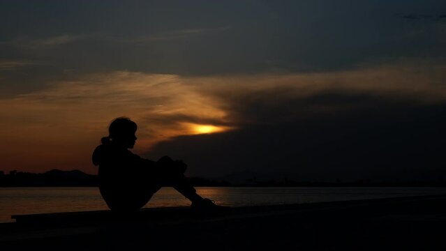 Depressed teen silhouette on sunset shore. A silhouette of depressed young girl sit in loneliness on the bay by the sea and think about something during sunset in summer.