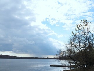 Cloudy sky above the smooth lake with leafless trees on the shore.