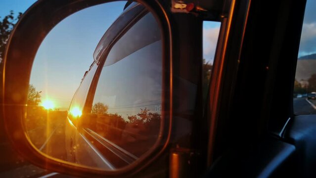 Closeup view of automobile mirror and man filming landscape in reflection on smartphone. He is driving on the car on asphalt road of Caucasus countryside at sunset and arriving to old wooden house