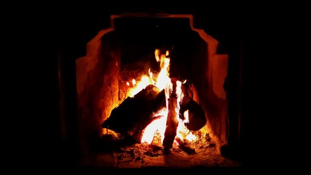 Burning timbers in vintage stone fireplace like in a cave filmed in darkness at night time. Closeup view of bonfire with hot logs and bright flames and sparks. Romantic evening spent next to campfire.