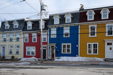 A street view of multiple colorful wooden homes with double hung windows. The closed glass windows have white trim. There's an orange, red, beige, and blue house with a cloudy sky in the background. 