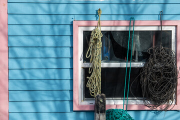 Multiple pieces of industrial nylon fishing rope hang from the trim around a pink and white colored closed glass window on a vibrant blue fishing shed. The wooden building has a horizontal clapboard.