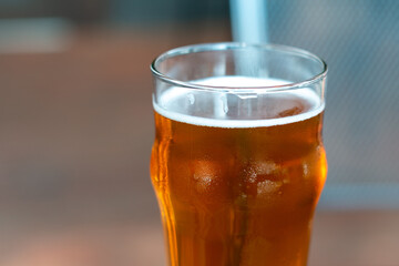 A clear pint drinking glass filled with cold froth from a lager ale. The Irish red ale pint sits on the edge of a metal patio table at a microbrewery. There are tables and chairs in the background.