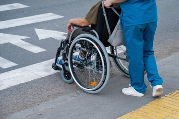 Obraz premium Rear view of a nurse helping an elderly woman in a wheelchair cross the road. 