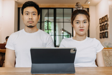 Straight faces of asian man and woman working with computer tablet in cafe.