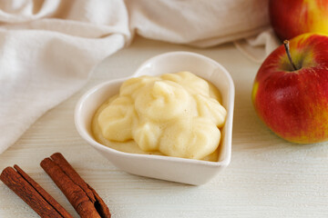 Fresh homemade applesauce, apple puree, mousse, sauce in heart shape bowl with cinnamon and apples on white table top side view Organic vegetarian food. Healthy eating