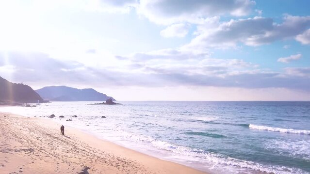 Tilt video on Sunset Beach where the sunlight pierces through the clouds backlighting the silhouette of a couple strolls along the shore of Sakurai Futamigaura's Meoto Iwa Couple Rock.