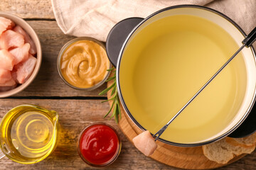 Fondue pot with oil, fork, raw meat pieces and other products on wooden table, flat lay