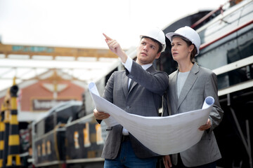 Young caucasian engineer man and woman in suit checking train looking blueprint in station, team...