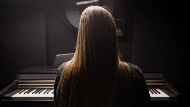 Long-haired girl composer sits with her back to the camera and plays the piano in recording studio