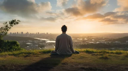 Foto op Plexiglas Meditation, harmony, life balance, and mindfulness concepts.A man sitting on a hill with grasses, meditating in silence, with the landscape of a city and bright morning sky. © KikkyCNX