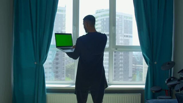 Rear view of a male student studying at home and using his laptop. Media. Man standing by the window and looking at green chroma key screen.