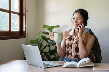A beautiful Asian woman is sipping coffee while talking on the phone at her desk in the home office.